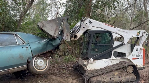 Skid steer removing an old junk car from a wooded property